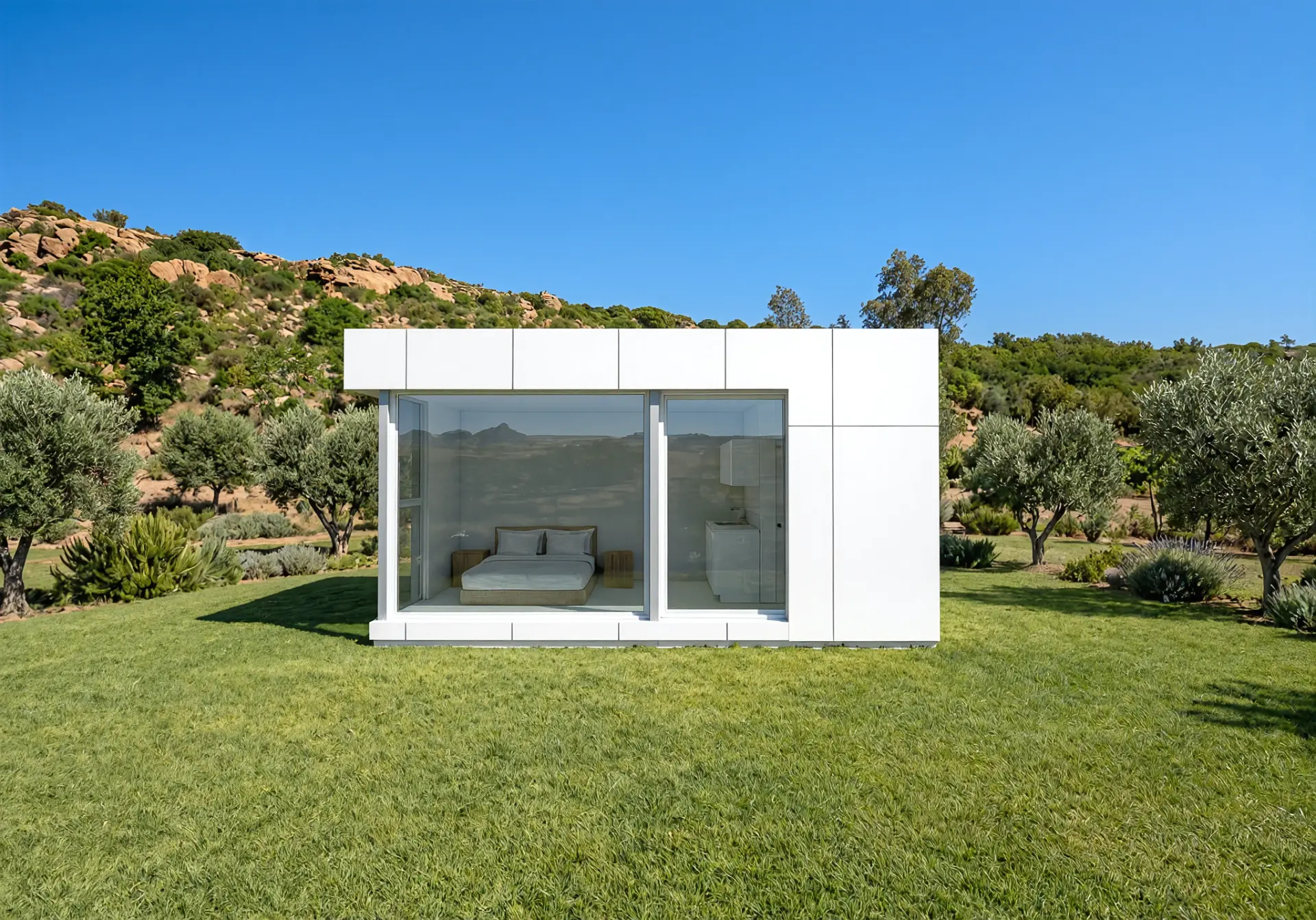 Modelo XS de frente: módulo blanco compacto con el dormitorio y la cocina de office visibles tras el vidrio, sobre césped entre olivos y una ladera rocosa